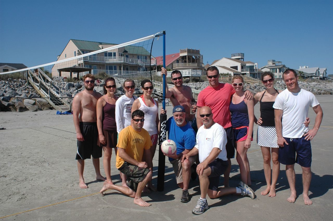 A group of people standing on the beach with a volleyball net.