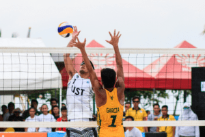 Two men on opposing teams playing volleyball outdoors with a crowd in the background